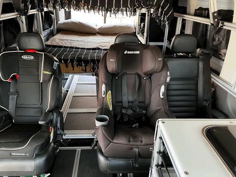 Modular RV rental interior featuring two child car seats & a bed for family adventures on the road.