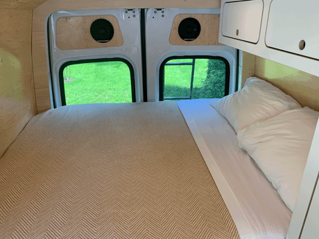 Cozy RV rental interior: a bed with herringbone blanket, wood walls, overhead storage, & views of nature through rear windows.