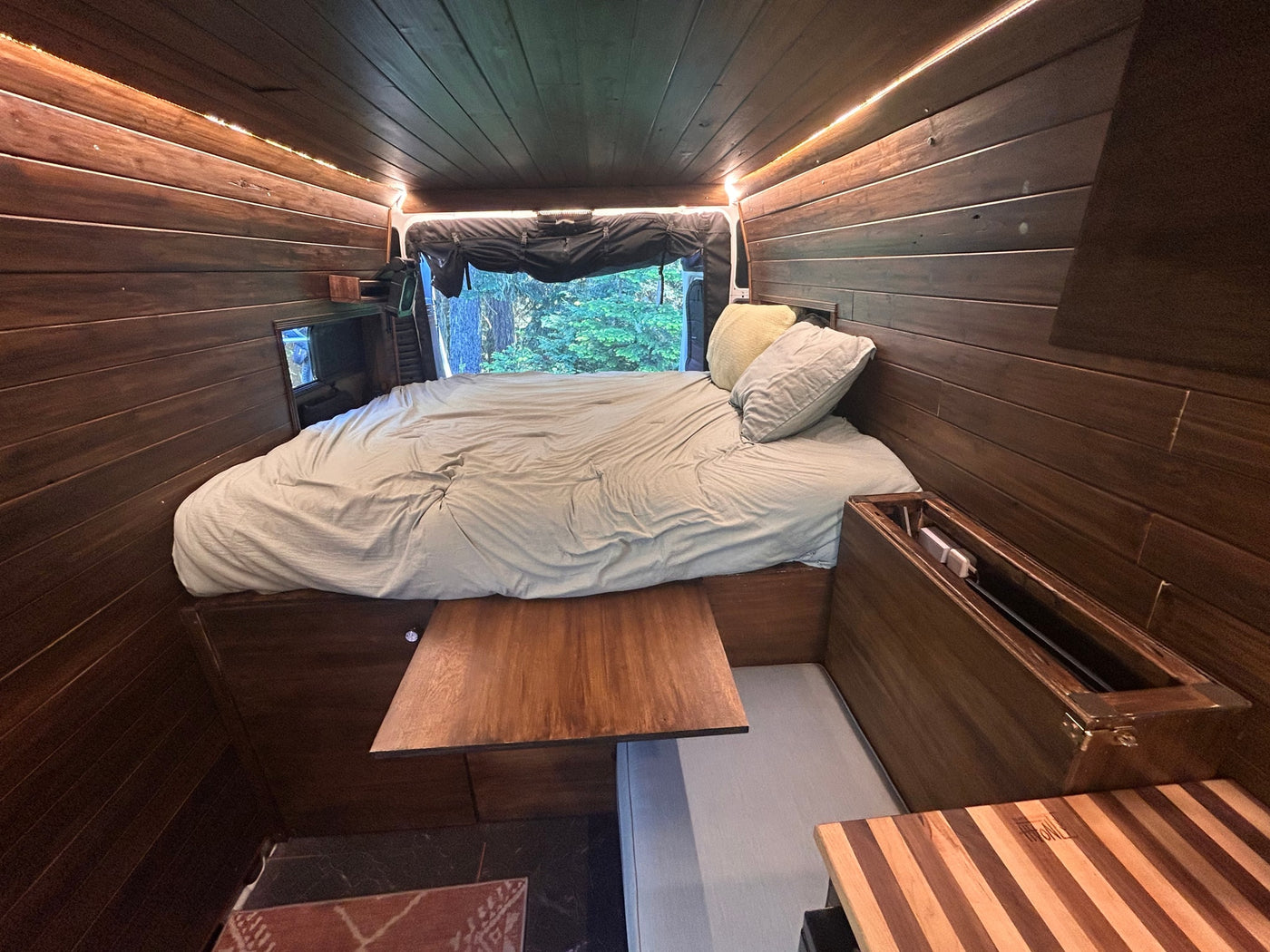 RV rental interior: cozy bed with light bedding, warm wood paneling, soft LED strip lights, & a forest window view. — photo 17