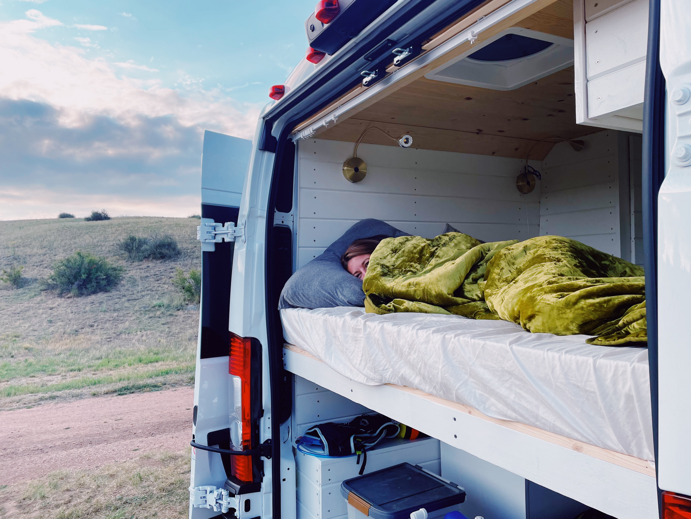 A person is sleeping under a green blanket in a white RV rental van, with the back doors open, revealing a grassy hillside.