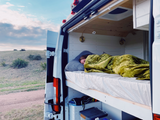 A person is sleeping under a green blanket in a white RV rental van, with the back doors open, revealing a grassy hillside.