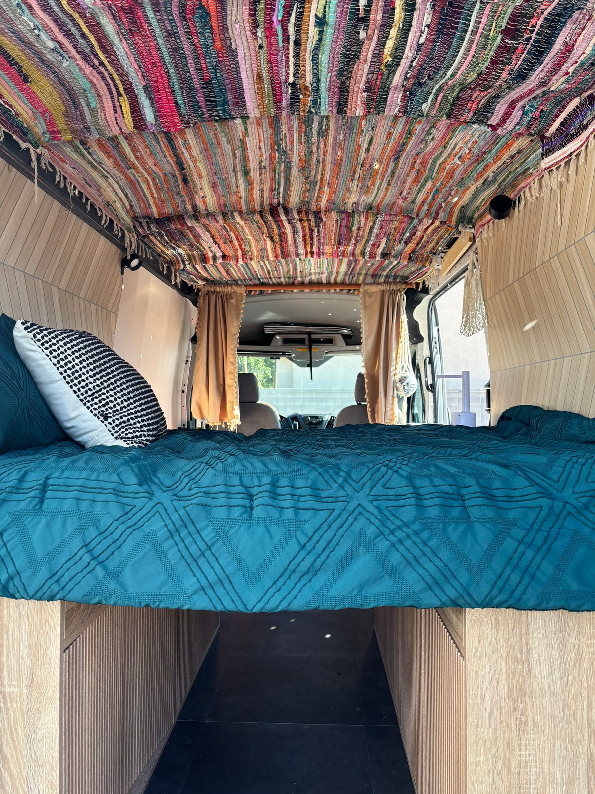 Colorful RV rental bedroom with a vibrant rag rug ceiling, warm wood paneling, and a cozy teal bed.