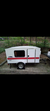 White Runaway Navigator teardrop camper, an ideal RV rental, parked amidst trees. Features a red stripe and small window.