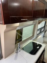Compact RV rental kitchen features dark wood cabinets, a sleek sink, and a black glass cooktop next to a large window.
