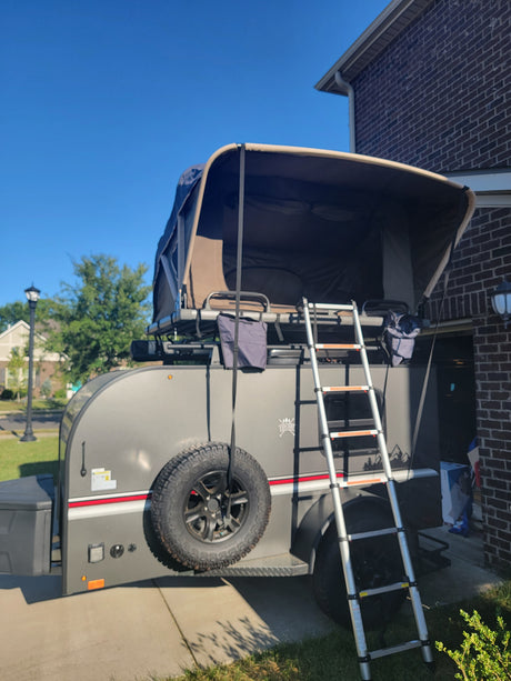 Grey pop-up camper for RV rental, featuring ladder & spare tire, parked on a sunny driveway.