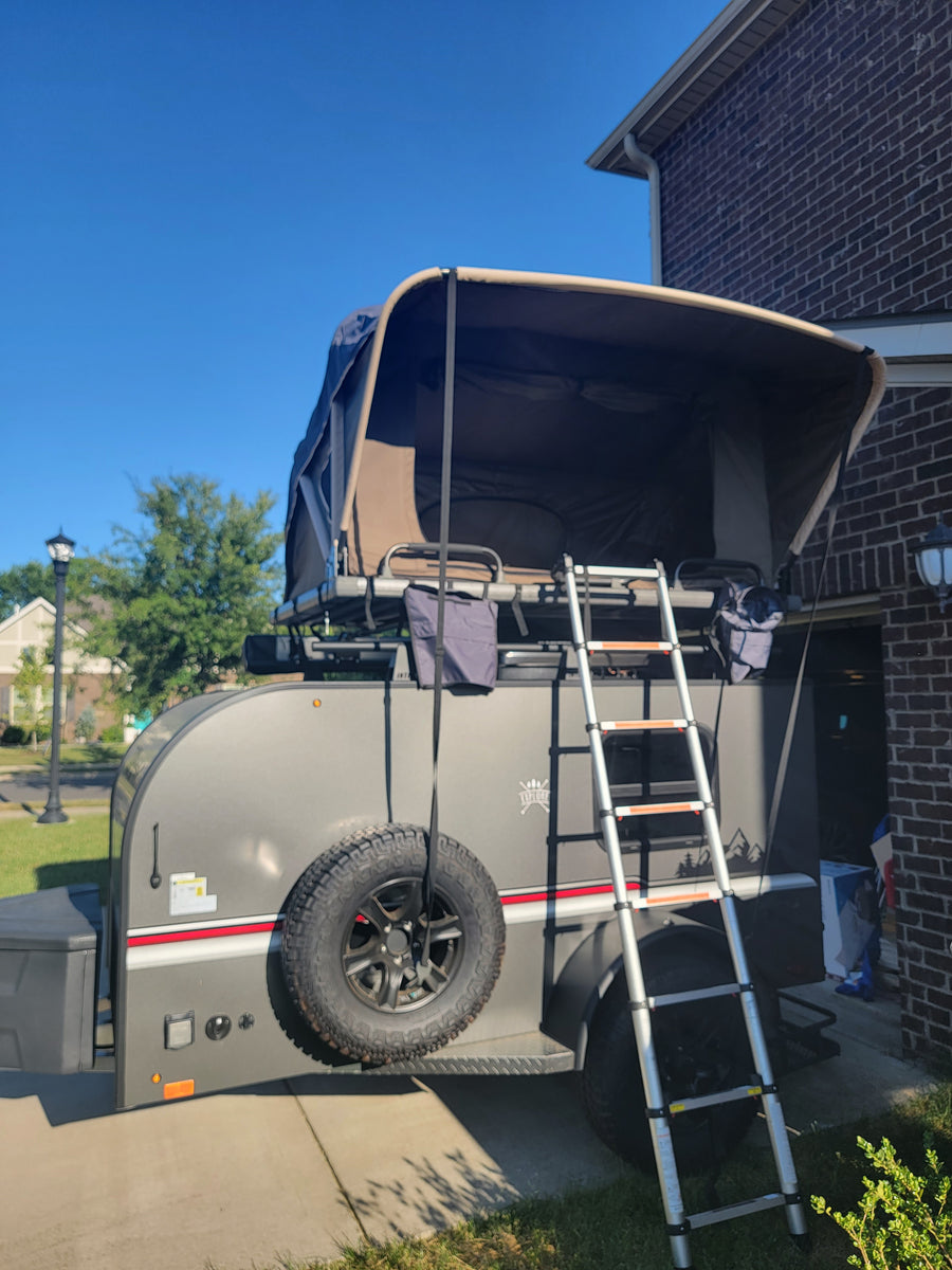 Grey pop-up camper for RV rental, featuring ladder & spare tire, parked on a sunny driveway.