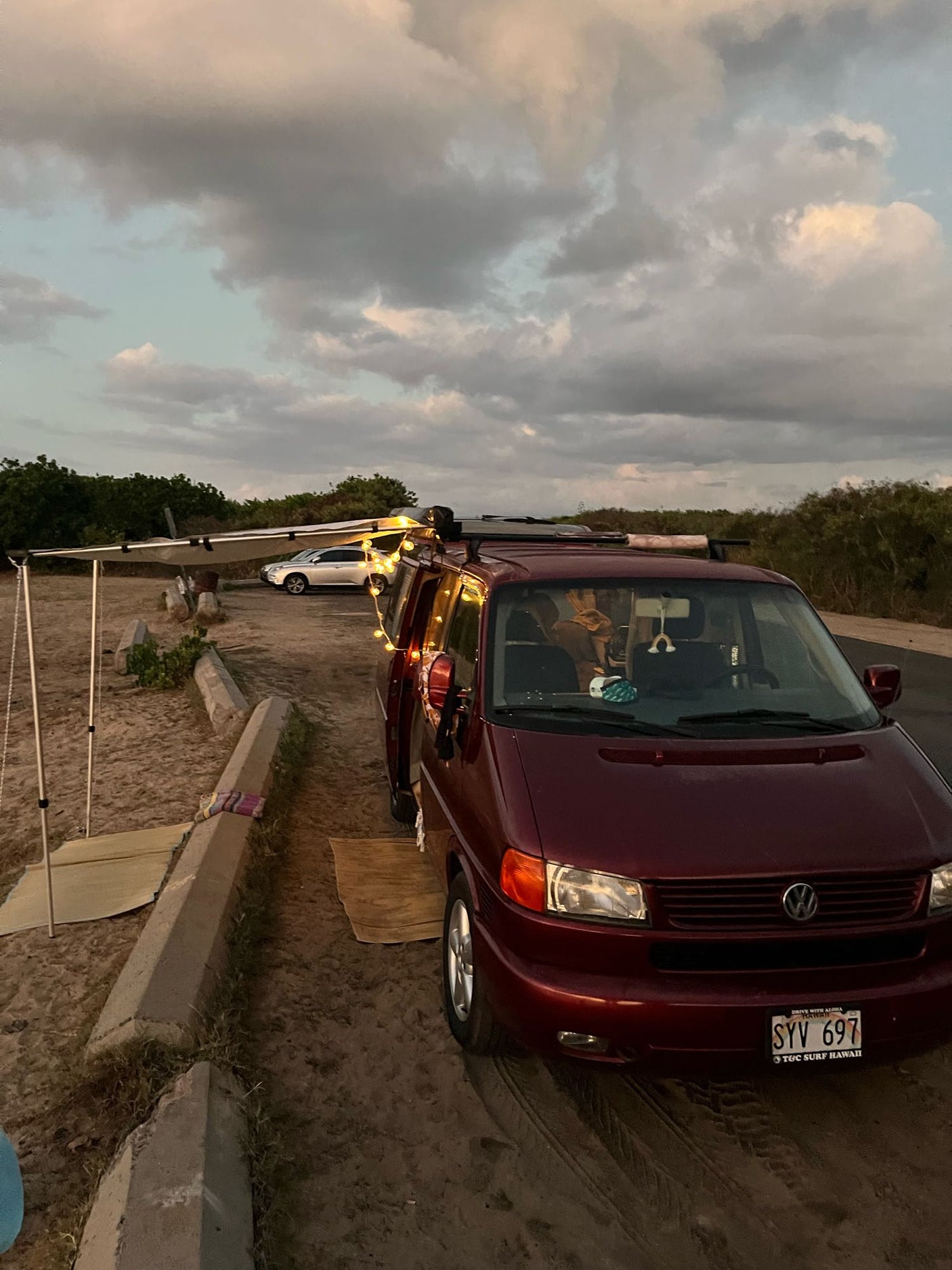 Evening setup with an RV rental: a burgundy VW camper van with an extended awning and string lights by a sandy road.