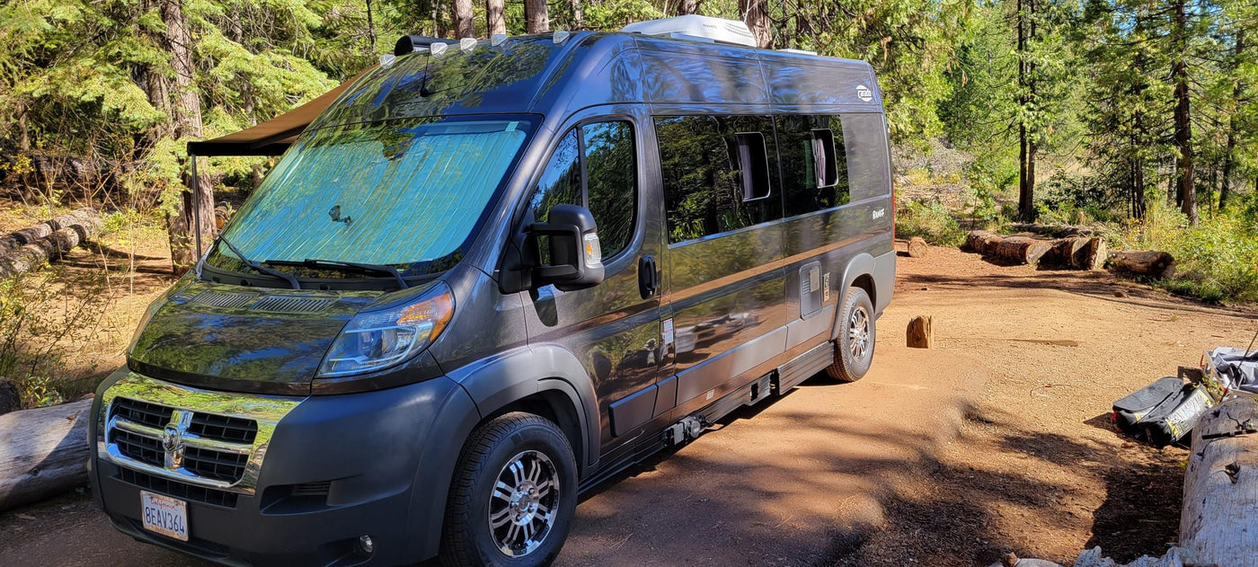 Dark gray camper van, possibly an RV rental, at a sunny wooded campsite with trees and logs. — photo 14