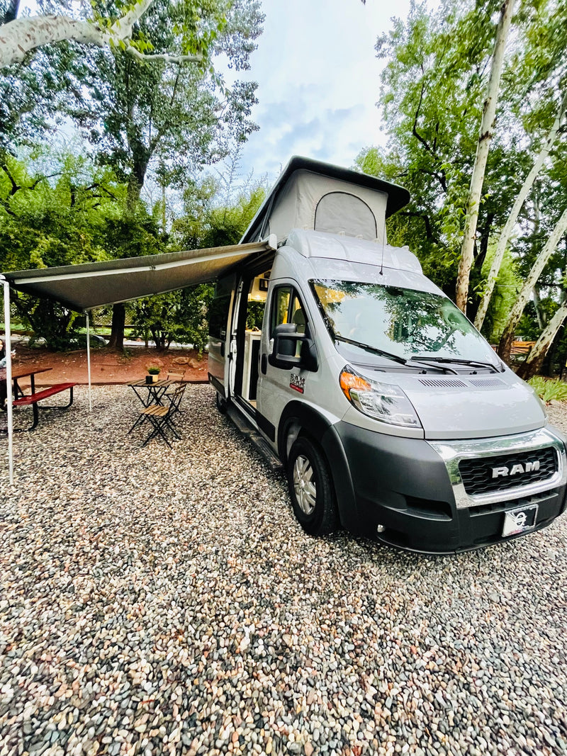 Gray **rv rental** campervan with pop-top & extended awning set up at a gravel campsite amidst lush trees. Perfect for outdoor adventure.|Silver Ram ProMaster camper van RV rental parked on gravel. Surrounded by lush trees — photo 1
