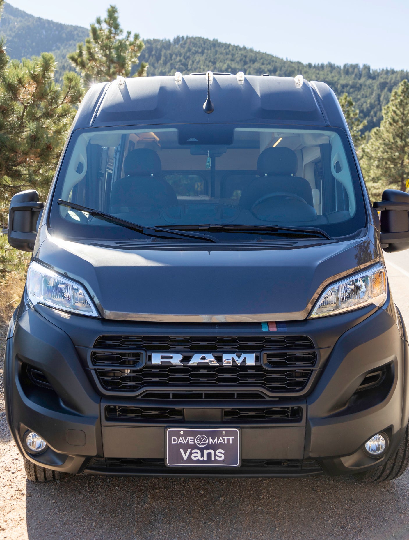 Front view of a dark gray Ram ProMaster campervan, likely an RV rental, parked on a dirt road in the mountains.
