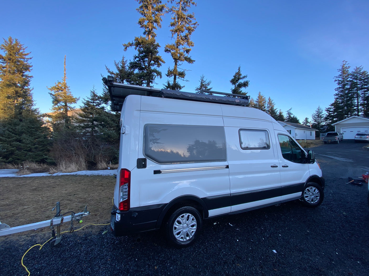 White RV rental camper van parked on gravel, with tall pines under a blue sky, trailer nearby. Perfect for outdoor adventure.