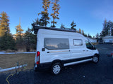 White RV rental camper van parked on gravel, with tall pines under a blue sky, trailer nearby. Perfect for outdoor adventure.
