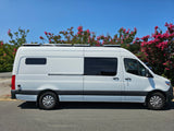A white camper van, ideal for RV rental, with roof panels and tinted windows, parked under a clear blue sky by pink flowering trees.