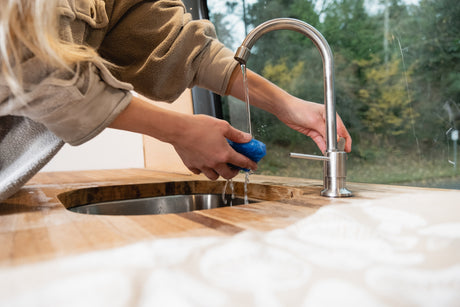 Hands washing a blue sponge under a faucet in an RV rental kitchen, featuring a wooden counter, sink, and forest views.