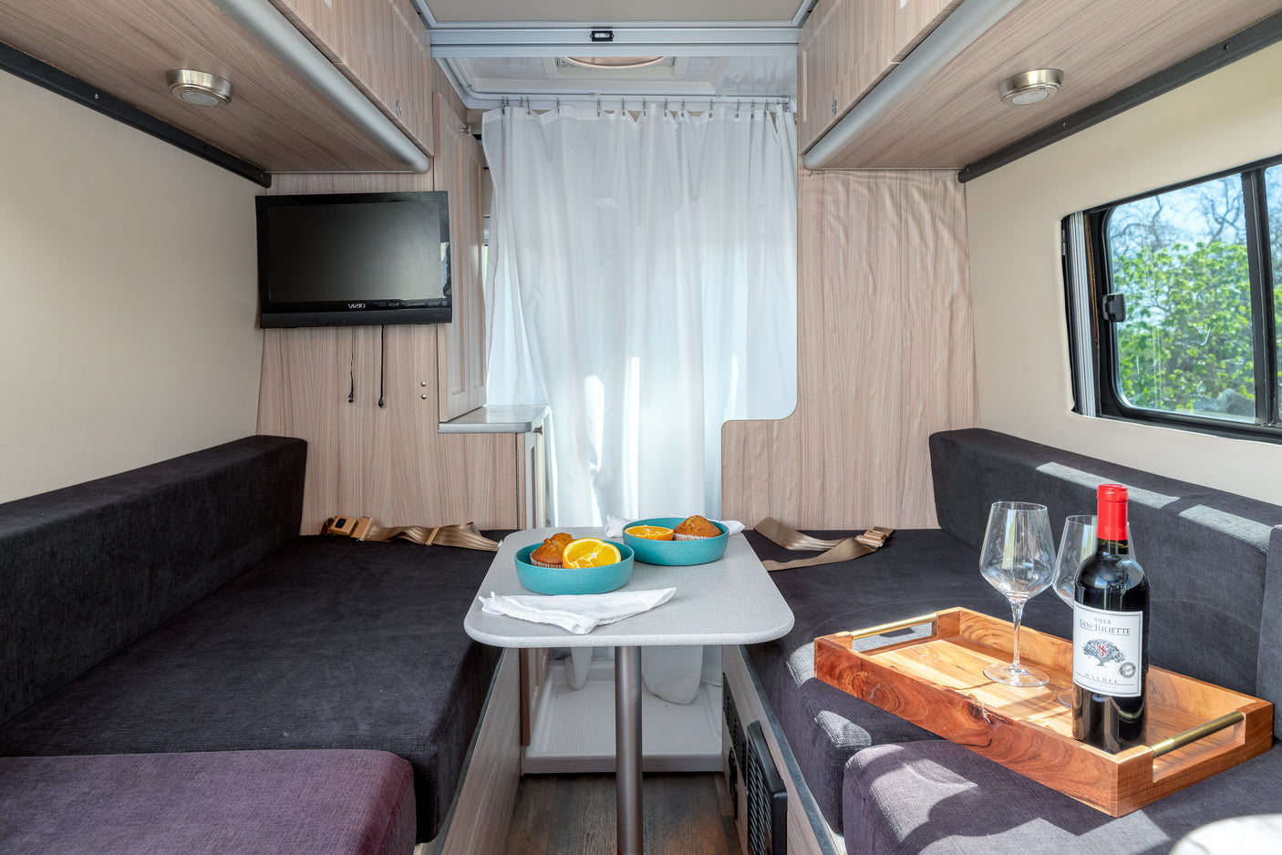 Cozy rv rental interior featuring facing benches, dining table with breakfast, TV, and wine for a comfy camper van experience.