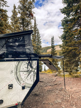 This RV rental camper trailer with pop-top tent sits lakeside, offering rugged forest and mountain views.