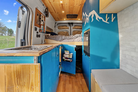 Inside this vibrant camper van RV rental: kitchen, bed, custom blue cabinetry with mountain art, and nature view.