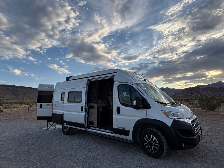 An RV rental (Winnebago Solis camper van) parked with doors open in a desert mountain landscape, light breaks through clouds.