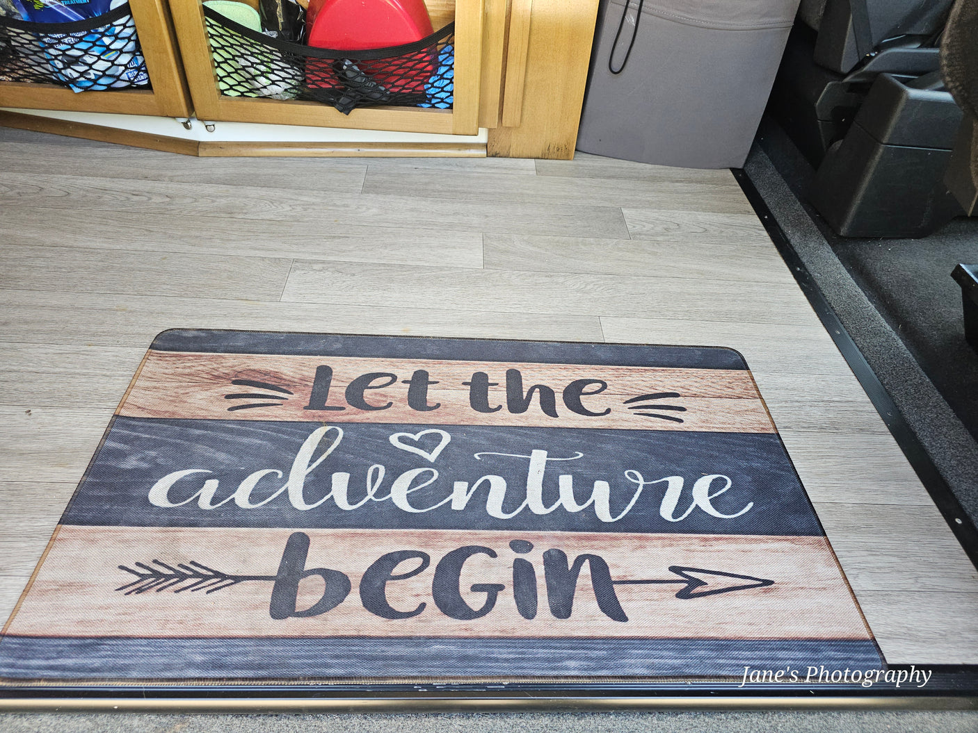 "Let the adventure begin" mat on a light wood-look floor inside an RV rental, with storage nets and a seat.