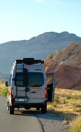 A grey Winnebago Revel RV rental van parked on a desert road with its door open, set against mountains and striking red rock formations.