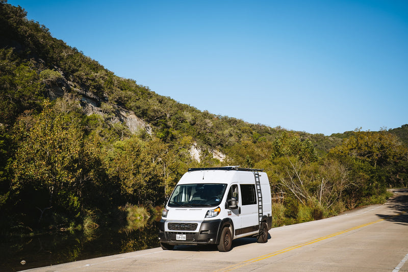 A white rv rental camper van is parked on a scenic road next to a calm river and a lush, tree-covered hill. — photo 13