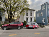 A red SUV tows a gray and red teardrop RV rental, parked on a street with houses and a tree. Note the wheel lock.