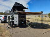 RV rental: Gray teardrop camper with roof-top tent & awning set up at a scenic campsite for camping & fishing.