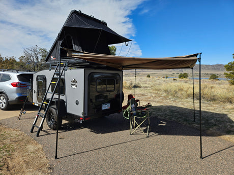 RV rental: Gray teardrop camper with roof-top tent & awning set up at a scenic campsite for camping & fishing.