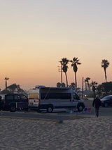 An RV rental and other vehicles are parked near a sandy area with palm trees silhouetted against a colorful sunset sky.