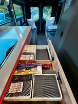 Organized kitchen drawer of an RV rental, featuring cooking supplies and remotes. A cozy living area is visible in the background.