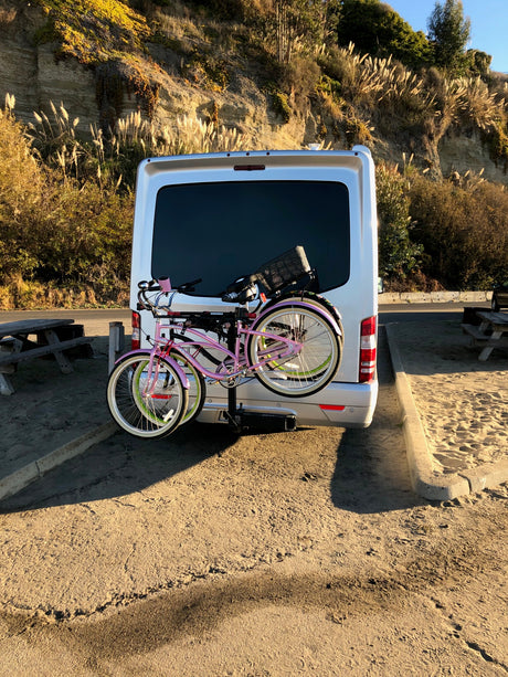 An RV rental van carries bikes on its rear rack, parked by a sunlit sandy cliff. Ready for scenic outdoor adventures.