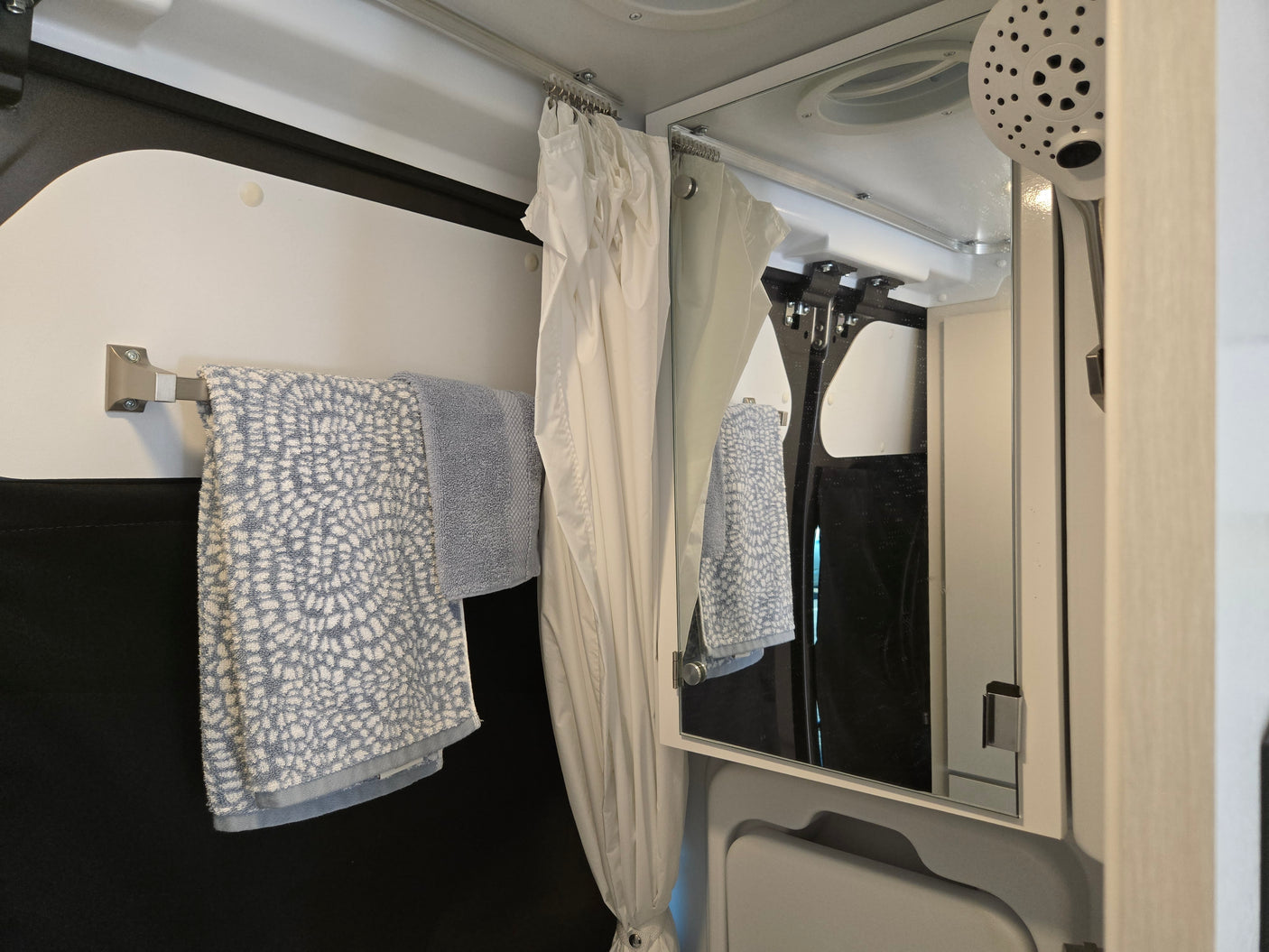 Inside an RV rental shower area: blue towels on a bar, a white shower curtain, and a mirror reflecting the space and a shower head.