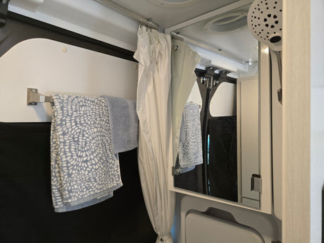Inside an RV rental shower area: blue towels on a bar, a white shower curtain, and a mirror reflecting the space and a shower head.
