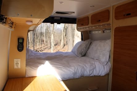 Inside an RV rental: a cozy bed with white linens, wooden cabinets, and a scenic forest view through the large window of this campervan.