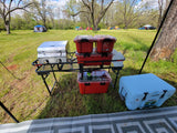 Portable camp kitchen with grill, red storage containers, and a blue cooler set up on a grassy field. Great for RV rental trips.