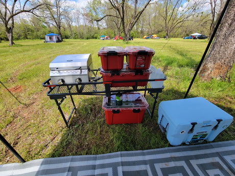 Portable camp kitchen with grill, red storage containers, and a blue cooler set up on a grassy field. Great for RV rental trips.