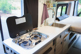 Compact rv rental interior with a functional kitchenette: two-burner stove, sink, and nearby bed.