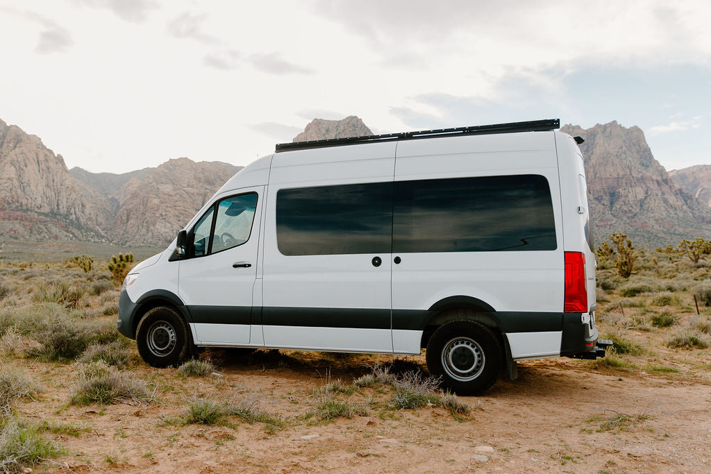 White RV rental camper van parked in a vast desert with rugged mountains under a cloudy sky.
