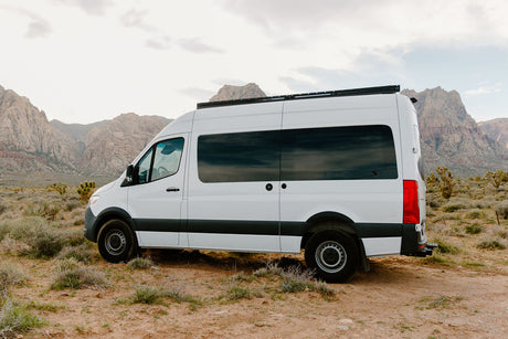 White RV rental camper van parked in a vast desert with rugged mountains under a cloudy sky.