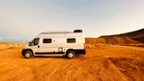 Off-grid RV rental adventure: A white camper van sits on a red desert track, surrounded by arid hills.