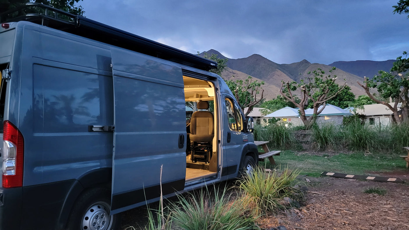 Grey RV rental campervan parked at a mountain campsite with an open door, revealing the cozy interior at dusk.
