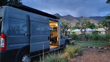 Grey RV rental campervan parked at a mountain campsite with an open door, revealing the cozy interior at dusk.