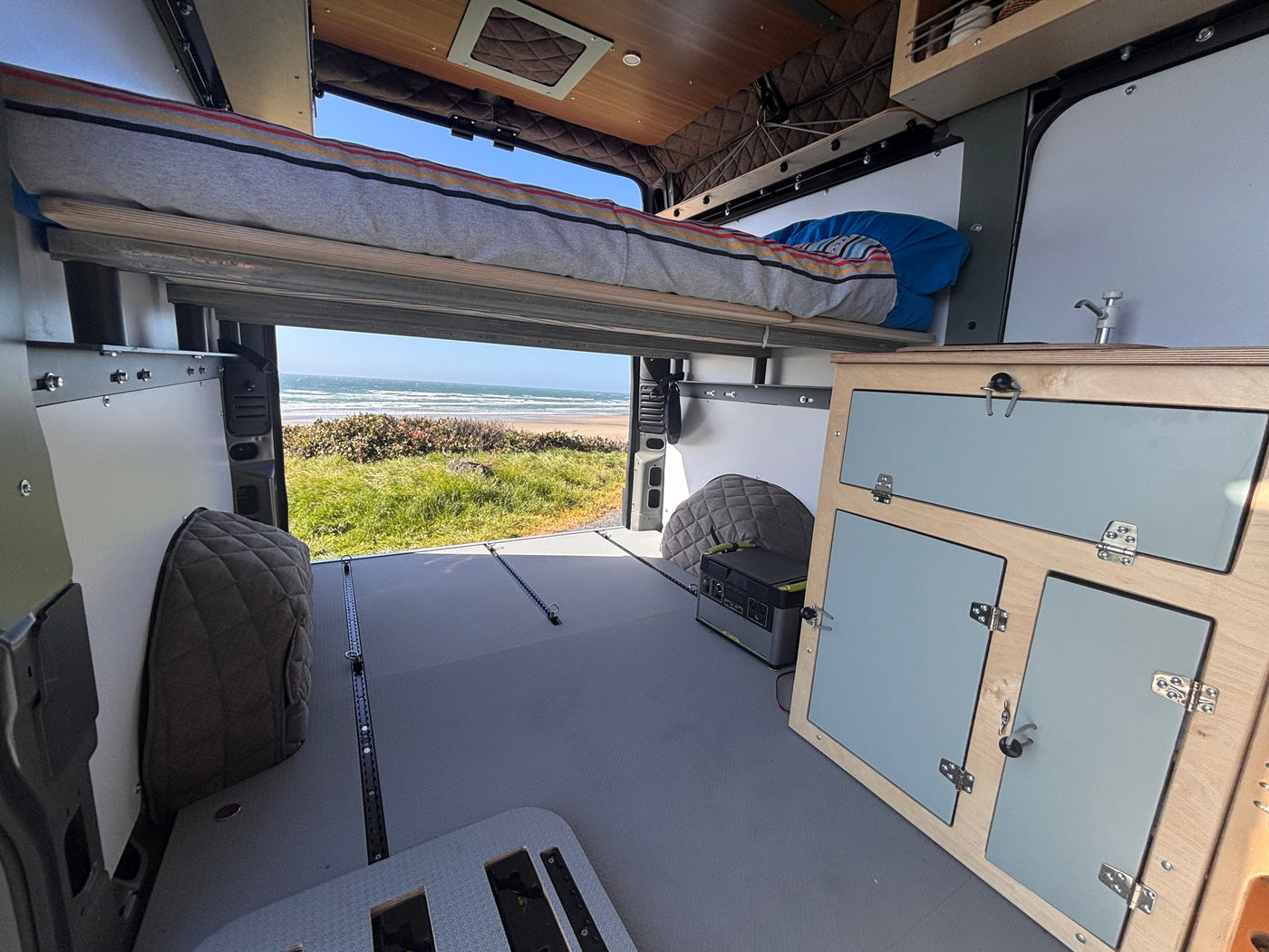 Enjoy this rv rental! Van interior with elevated bed, floor tracks, cabinet, and stunning ocean beach view. — photo 5