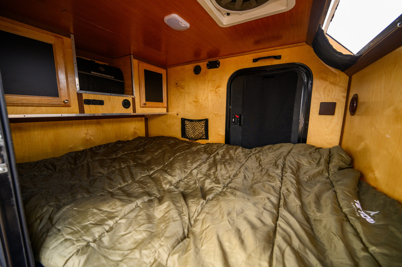 Inside a compact RV rental's cozy sleeping area, featuring a large olive green bed, wood interior, and functional built-ins.