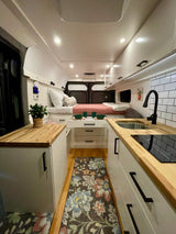 Bright RV rental interior: bed, sleek kitchen, white cabinets, wood counters & floor rug create a functional, cozy space.