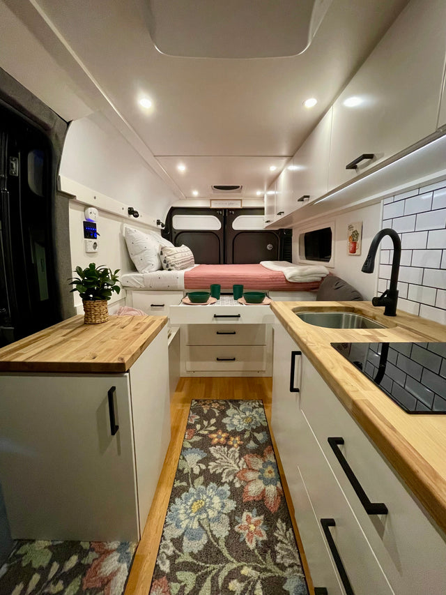Bright RV rental interior: bed, sleek kitchen, white cabinets, wood counters & floor rug create a functional, cozy space.