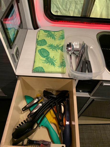 Inside an RV rental: a neat kitchen with a utensil-filled drawer, countertop cutlery, a pineapple towel, and controls.