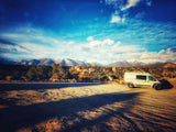 A white camper van (rv rental) parked in a sunlit, dusty landscape with long shadows, backed by snow-capped mountains and a blue sky.