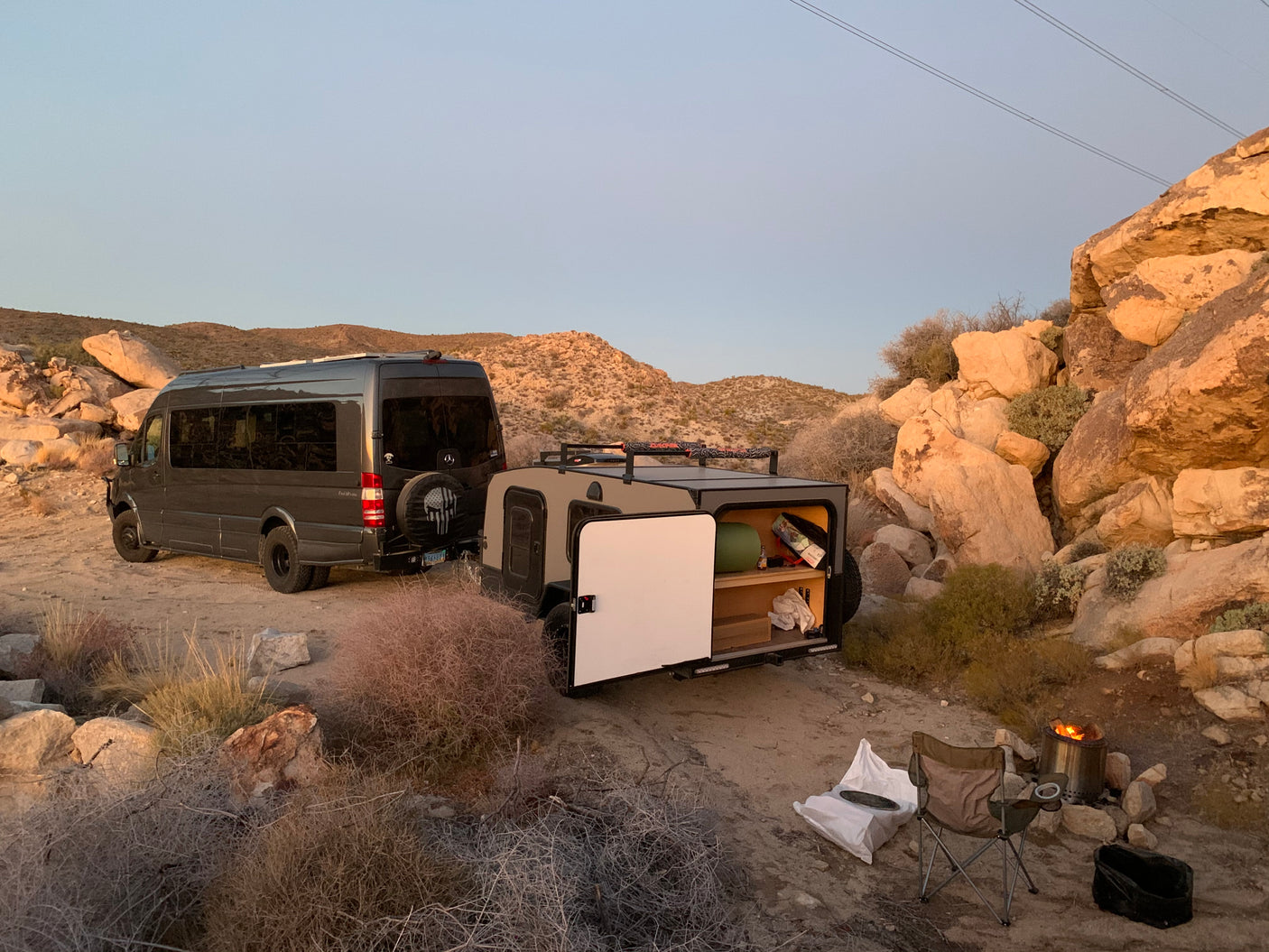 Perfect RV rental getaway: A dark grey camper van & tan trailer set up camp in a rocky desert, with a cozy fire pit.