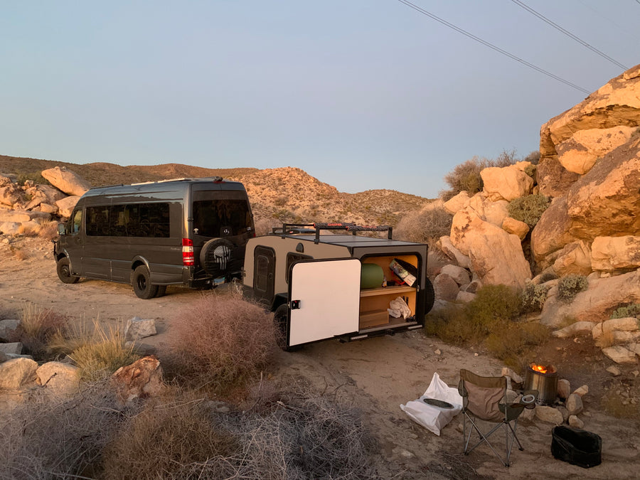 Perfect RV rental getaway: A dark grey camper van & tan trailer set up camp in a rocky desert, with a cozy fire pit.